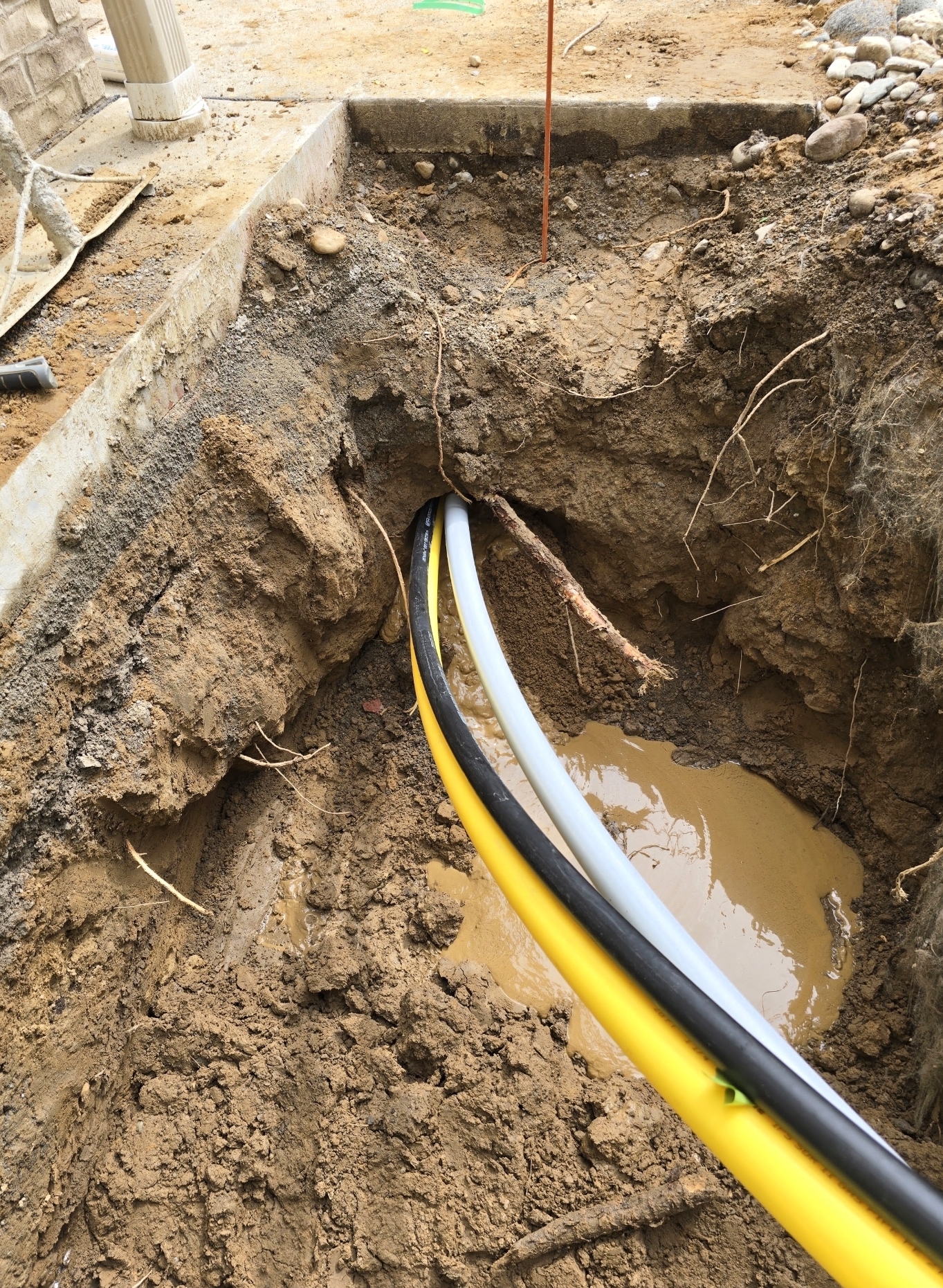 Yellow, black, and grey utility conduits emerging from a bore tunnel in an excavated access pit beside a building foundation