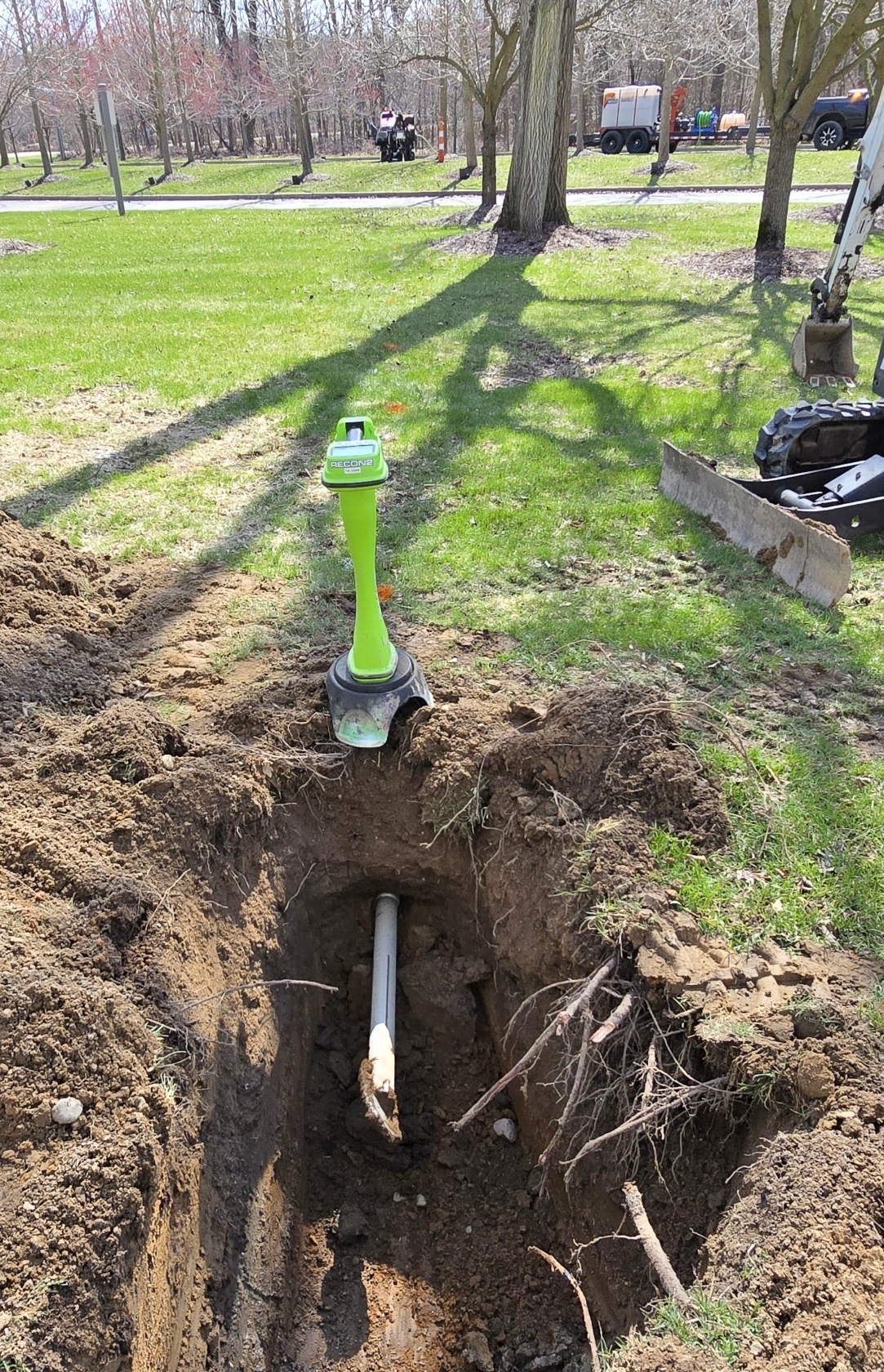 Directional drill head and pipe stem emerging through the soil wall of an excavated receiving pit, with a green locating receiver standing over the bore path
